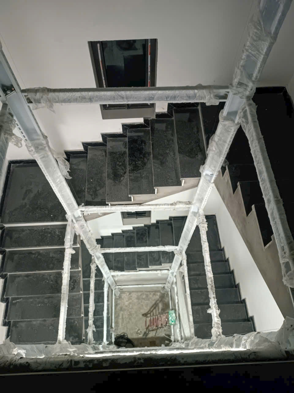 Cầu thang đá đen trong nhà đang thi công nhìn từ trên xuống, lan can bọc nilon bảo vệ